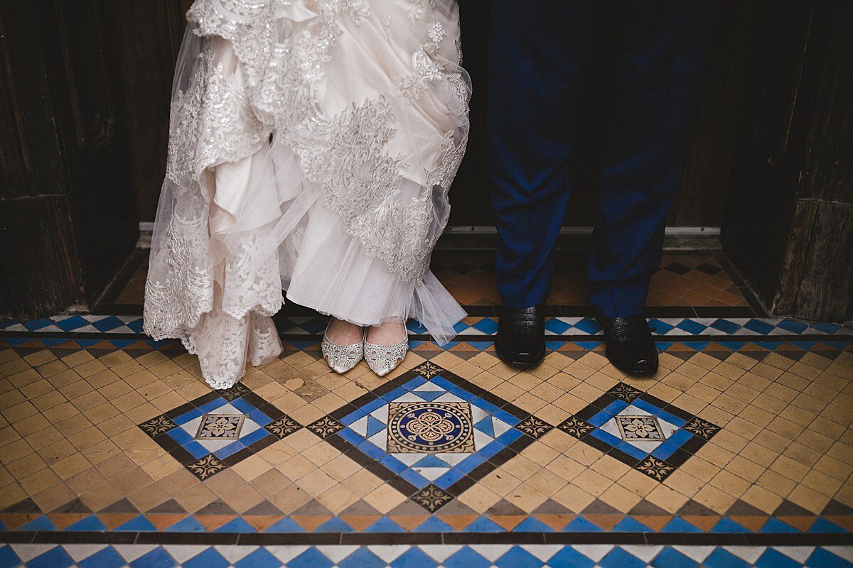 The bride is wearing a ruffled layered tulle and lace wedding dress and beaded heels. The groom is wearing black pants and black dress shoes. They stand in front of a yellow, beige, blue and ivory mosaic floor detail.