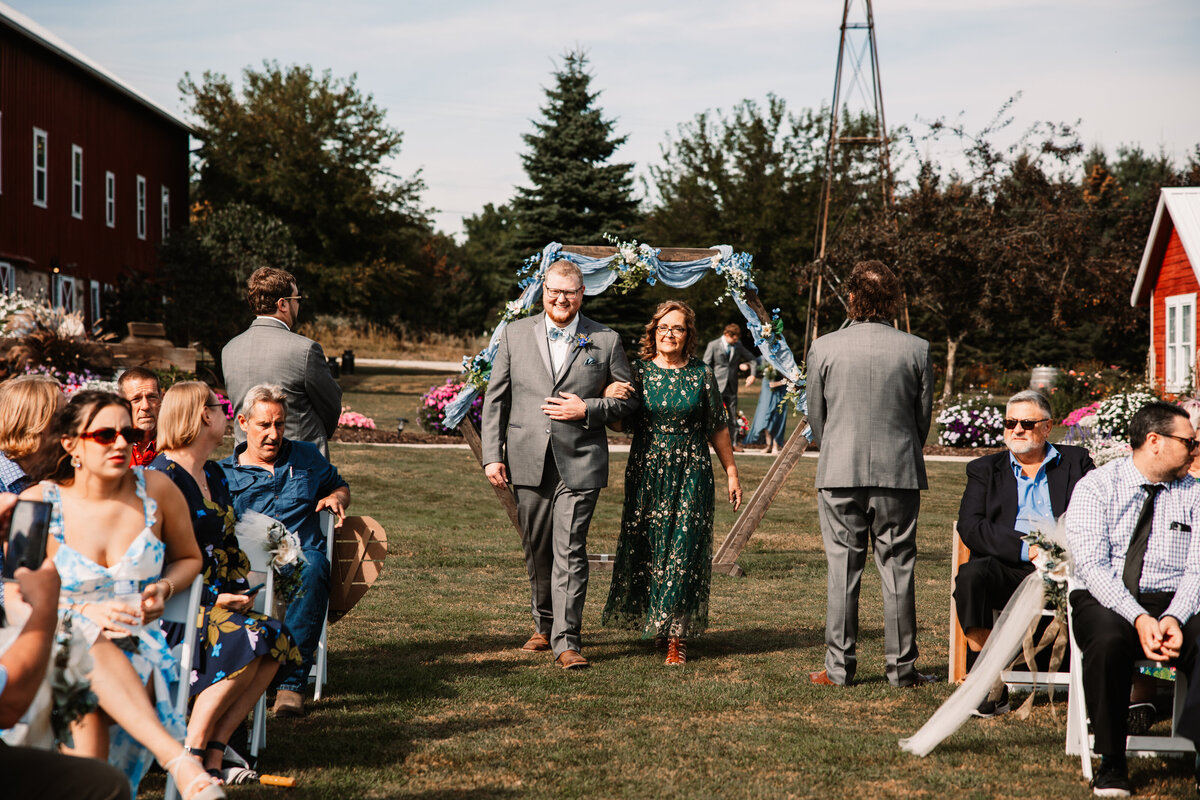 A groom in a grey suit walks down the aisle with his mother holding his arm