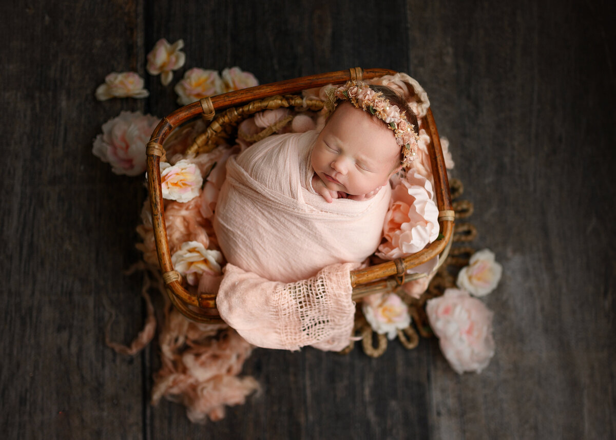 sweet newborn girl in pink swaddle posed on a bamboo basket  with soft pink floral headband
