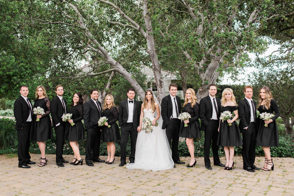 A bride and groom stand with their wedding party dressed in chic black attire, posing under lush green trees for a timeless and elegant wedding portrait.