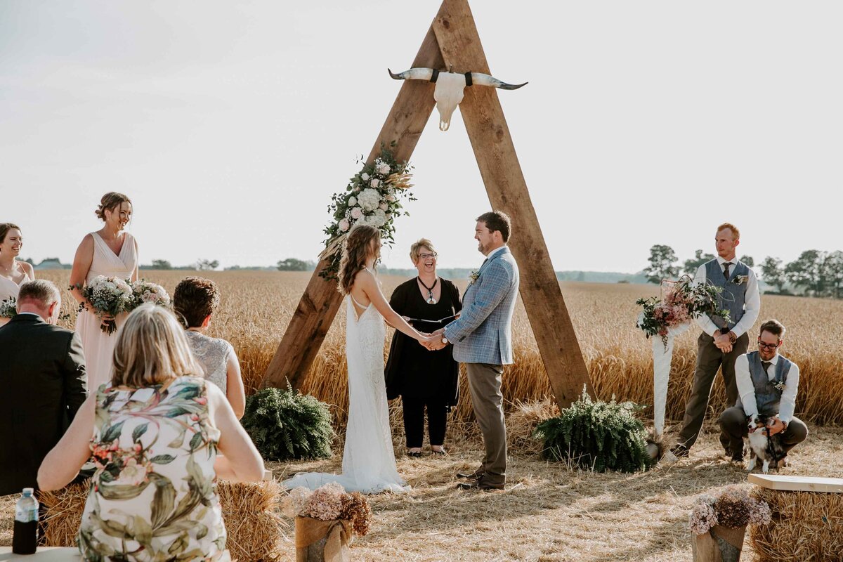 Bride and groom exchange vows at a rustic farm wedding in Exeter, Ontario. Bride and groom are holding hands with a wood arch behind them covered in white flowers and a bullhorn. Guests are sitting on haybales.
