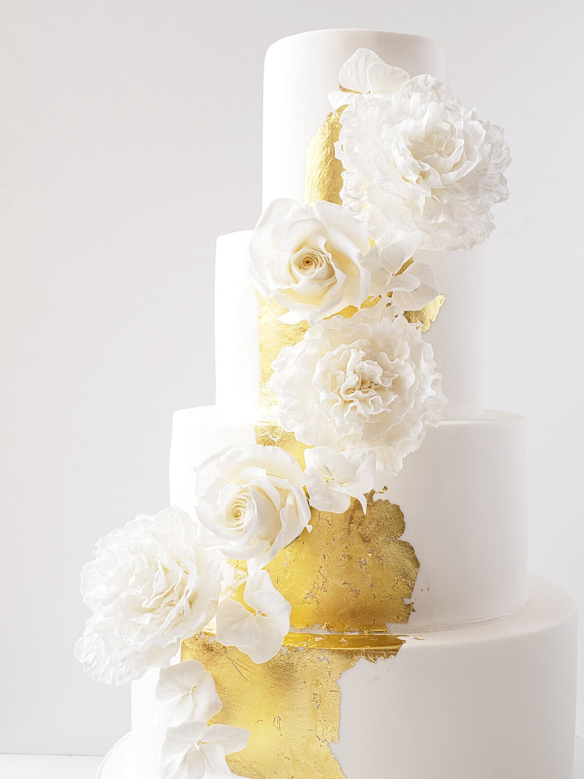 Elegant 3 tier white wedding cake with gold foil details and white roses cascading, created by Brianne Gabrielle Cakes,  elegant cakes & desserts in Edmonton, AB, featured on the Brontë Bride Vendor Guide.
