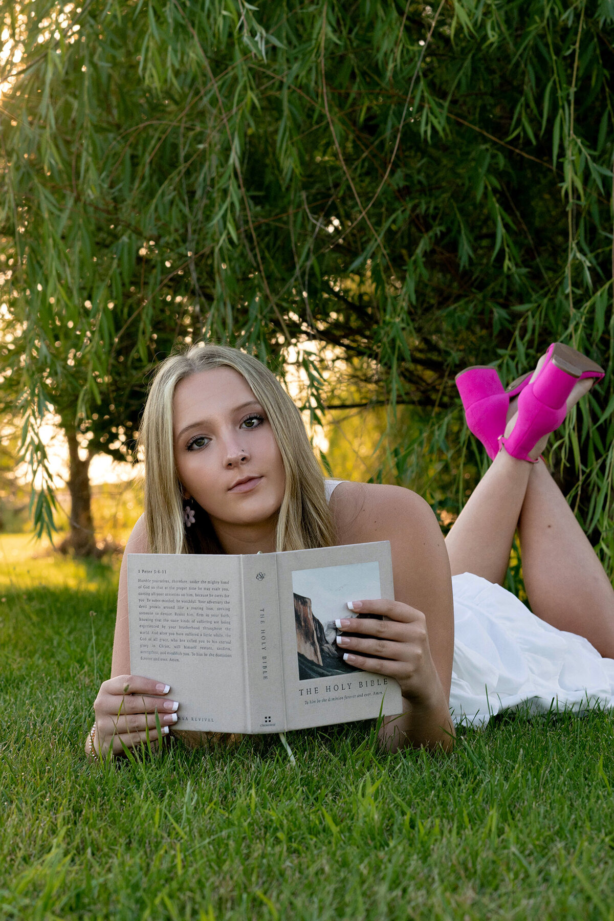A high school senior lays in the grass under a willow tree at sunset while reading the bible in a white dress and pink shoes