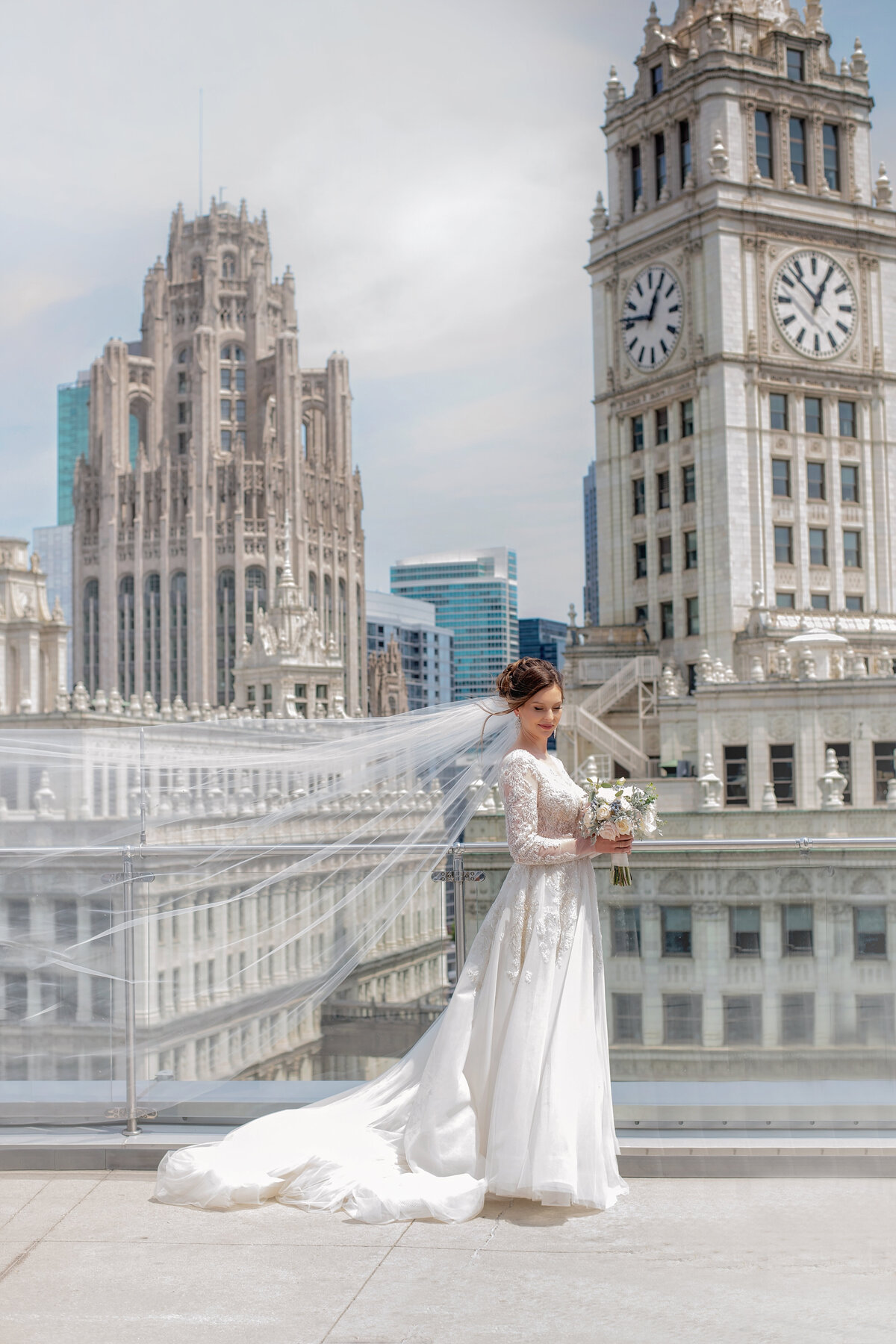 beautiful bride in stunning white wedding dress with long cathedral  veil surrounded by skyscrapers on Trump hotel rooftop in Chicago.