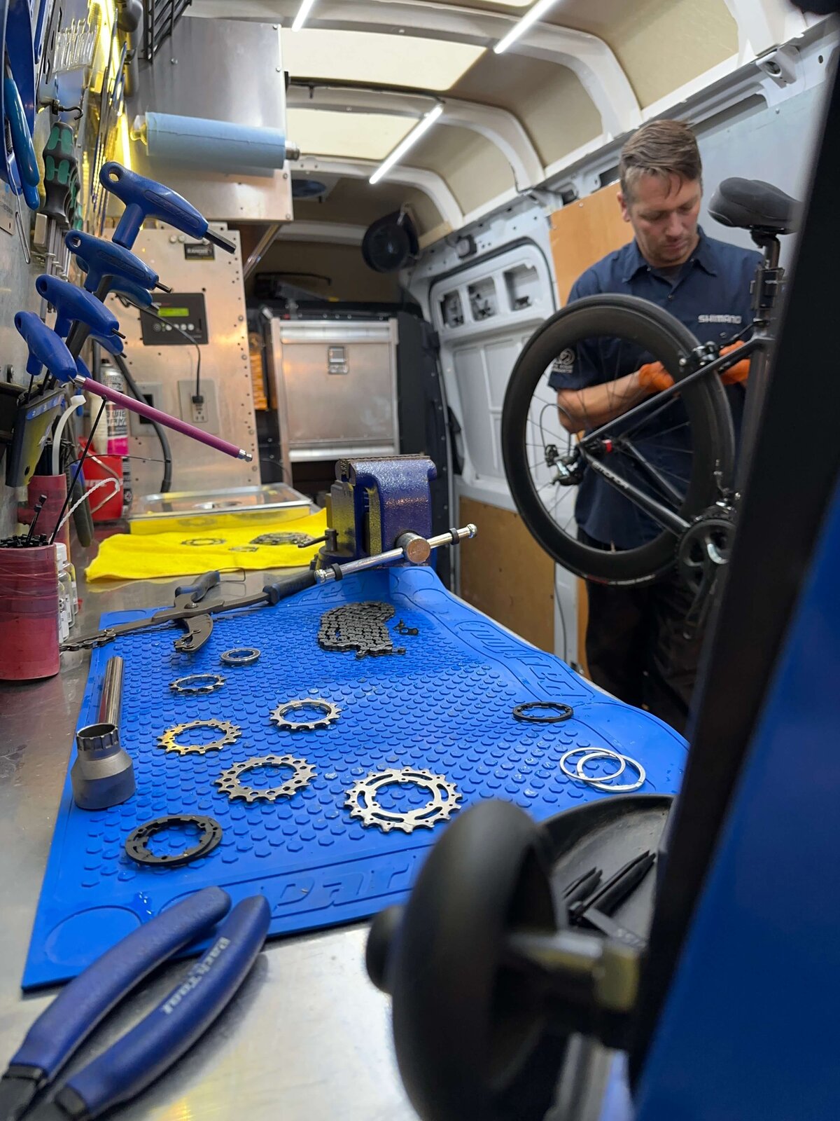 A person is repairing a bicycle inside a workshop van. Tools and gear components are neatly organized on a blue mat on a workbench. Various maintenance tools hang on the walls, and the van appears well-equipped for mobile bike servicing