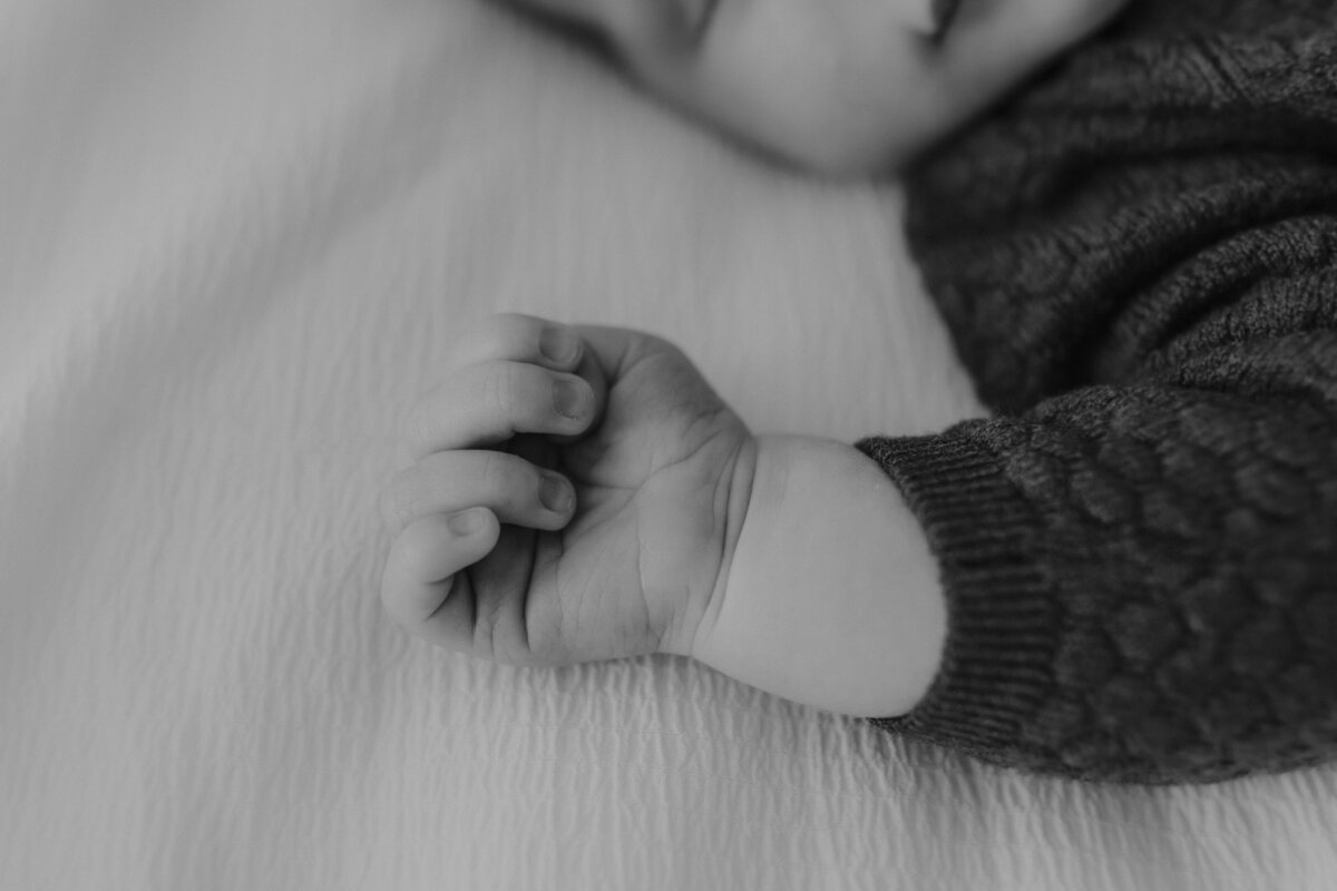 Black and white image of a baby's hand resting on a soft surface. The baby is wearing a knitted sweater.