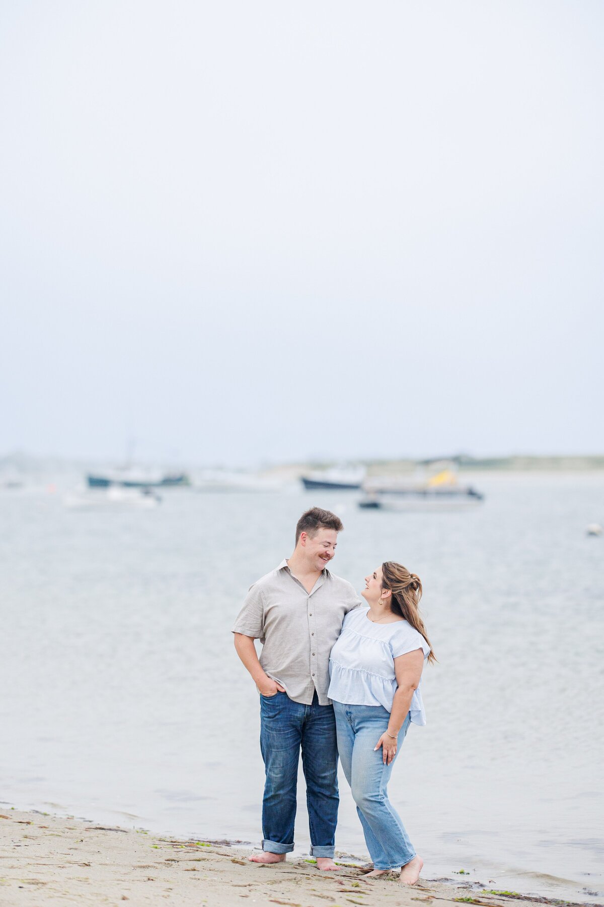 Couple standing barefoot on the shoreline, smiling at each other with hands in pockets and anchored boats in the blurred background at Chatham Bars Inn in Chatham, Massachusetts — representing the portfolio of a Cape Cod engagement photographer.