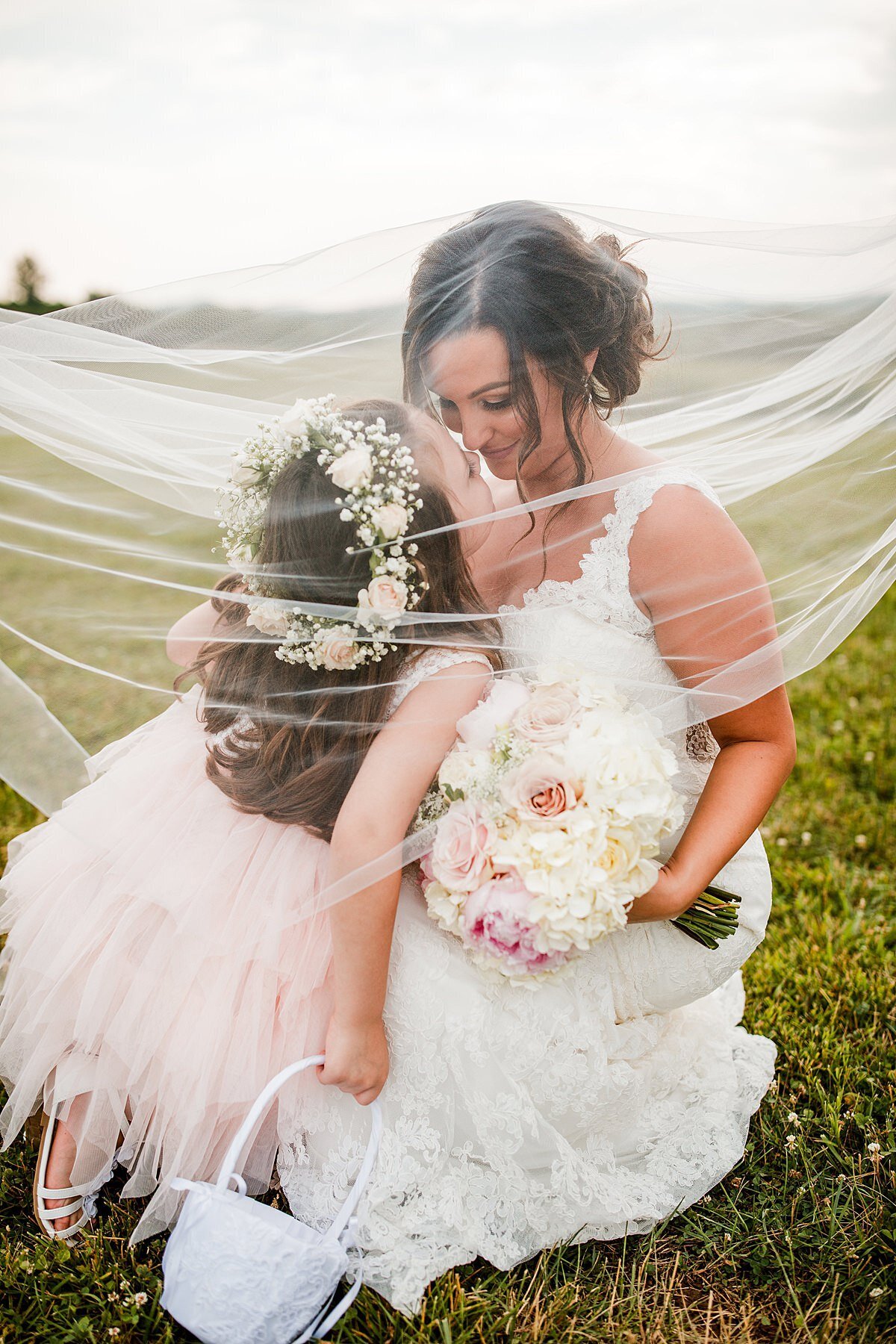 A bride and flower girl cuddle in a field underneath the bridal veil. The flower girl is wearing a very light pink ruffled dress with white sandals and a carnation and babys breath flower crown while holding a white satin basket of flower petals. The bride is wearing a white lace wedding dress and is holding a white round bouquet of hydrangea, white roses and blush pink roses at White Dove Barn for her Nashville Wedding.