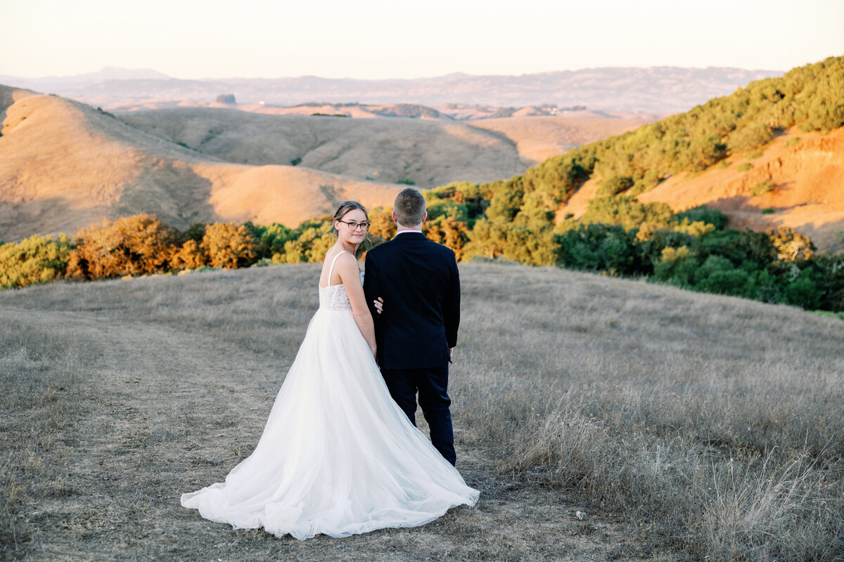 wedding photographer bay area photographs wedding pictures with groom looking at the hills in Petaluma with the bride holding her grooms arm and looking over her shoulder to the camera with the sun shining over the hills at sunset