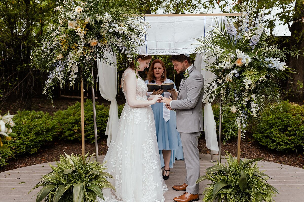 Redheasded bride wearing a flowing white lace wedding dress holds hands with the groom wearing a white kippot and light grey suit under the chuppah. The officiant wearing a light blue dress stands under the chuppah decorated with the groom's tallit and large sprays of fern, greenery, white and blush flowers.