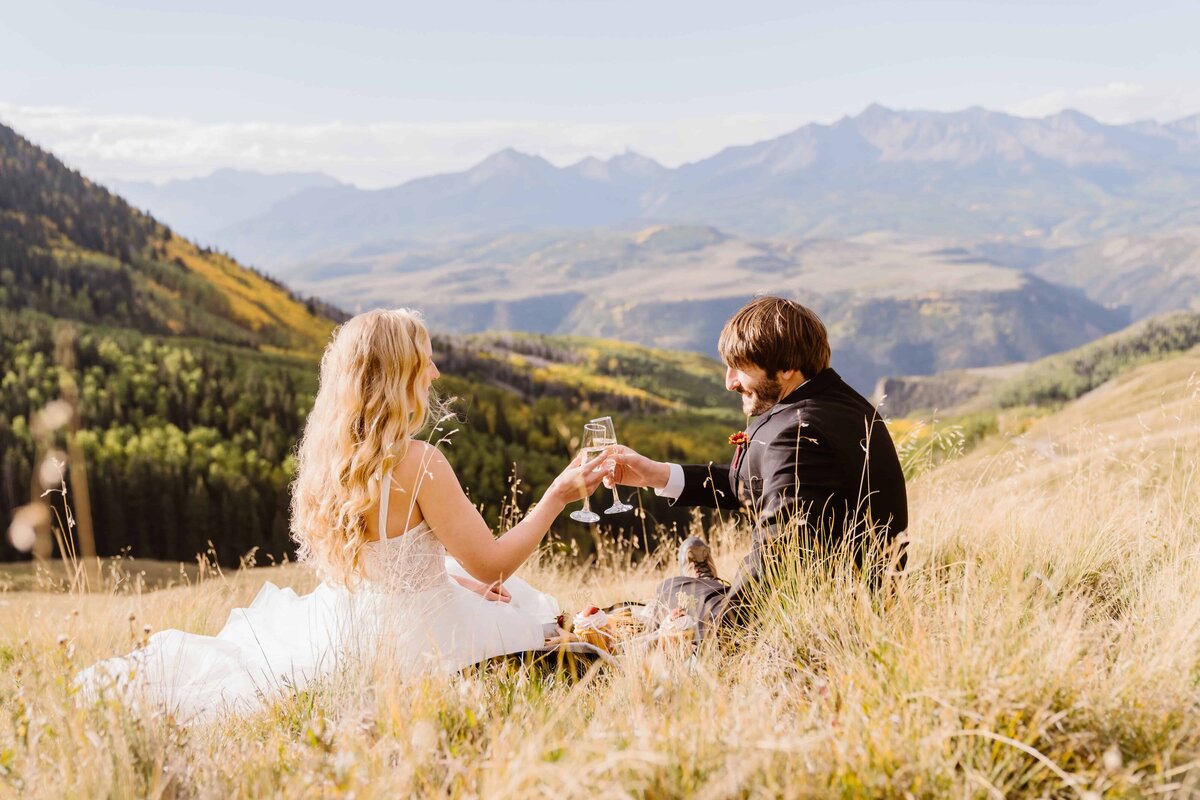 Couple in wedding attire sharing a champagne toast on a picnic blanket overlooking the Telluride mountains.