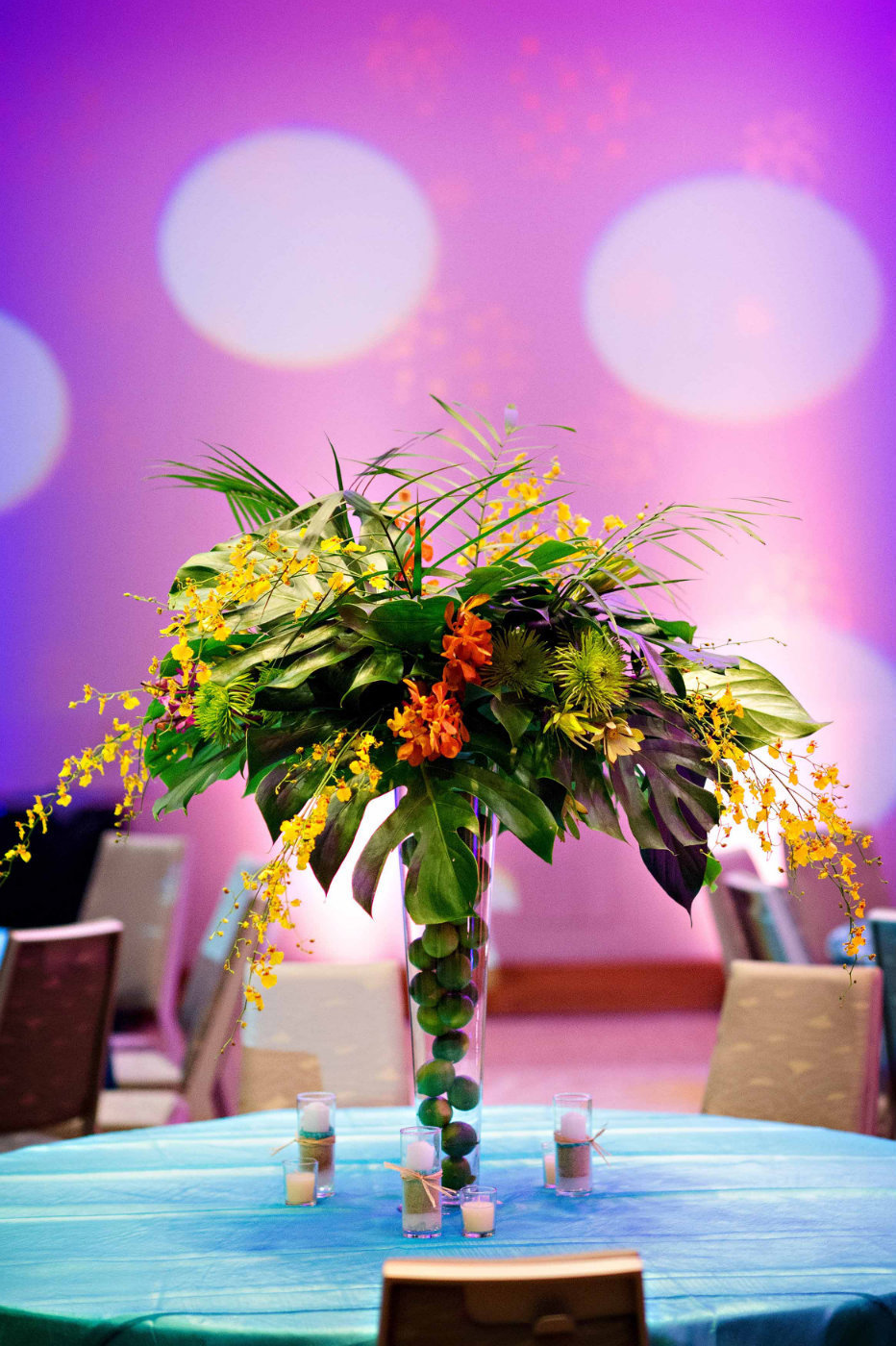 tall centerpiece with tropical leaves, purple orchids, in vase filled with limes on turquoise linen