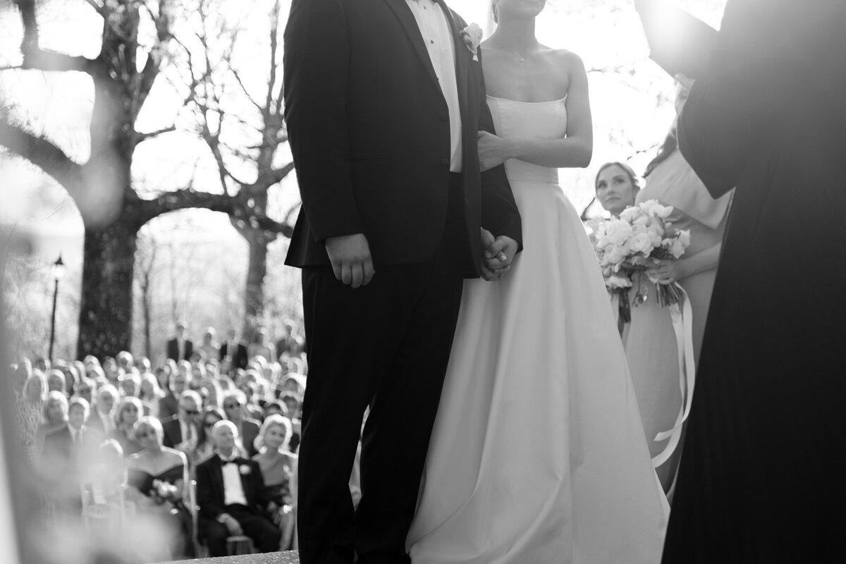 Black and white photo of the bride and groom standing together and holding hands during their outdoor wedding ceremony at Burritt on the Mountain in Huntsville, Alabama, photographed by Perry Hancock.