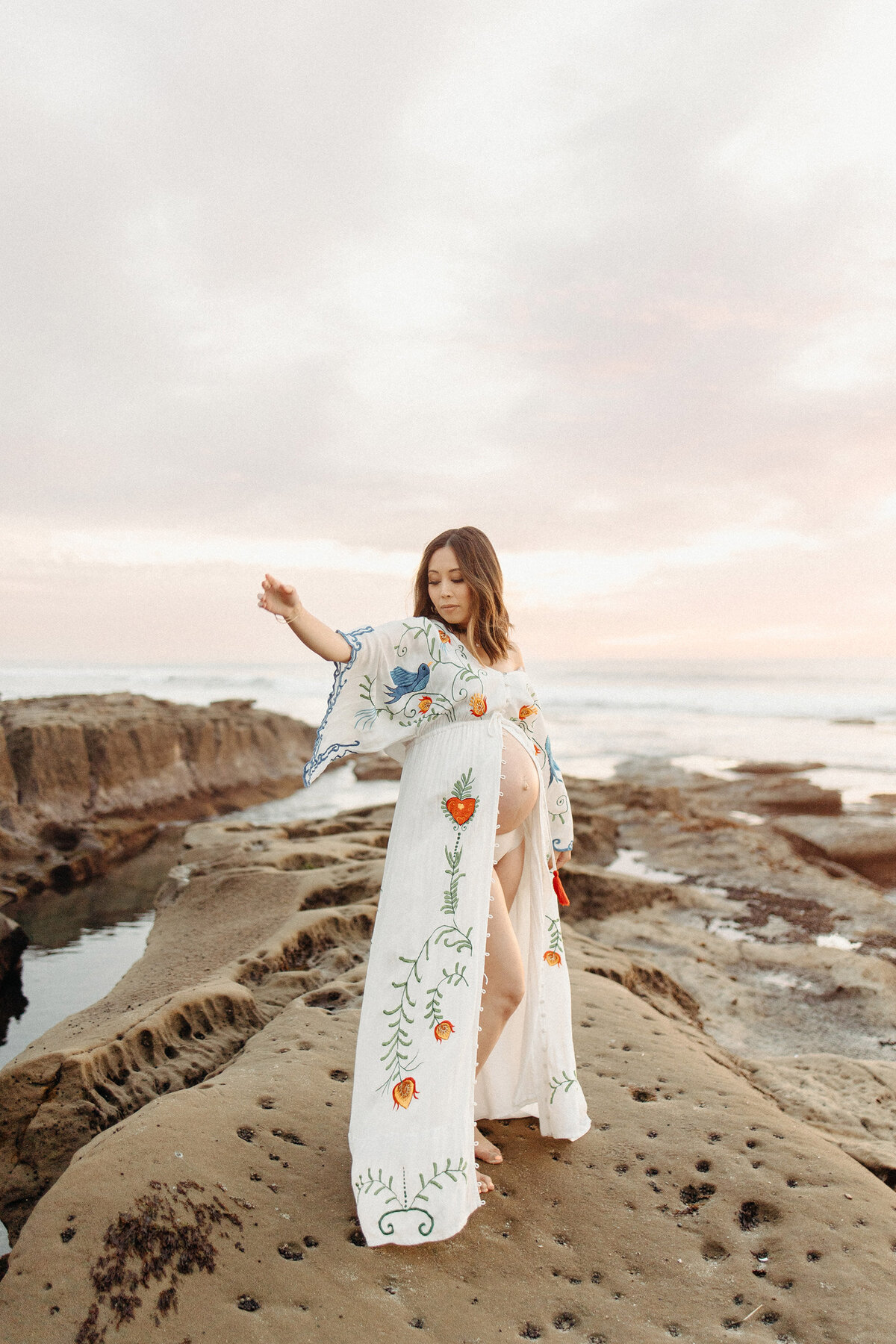 Elegant maternity portrait of a mother posing barefoot on rocky cliffs at La Jolla beach during sunset. Captured by San Diego Family Photographer Yuka Schad.