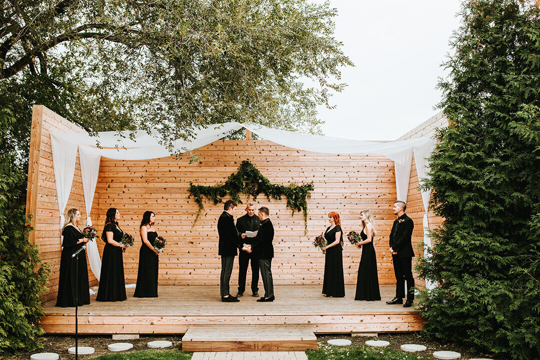 Two grooms wearing black tuxedos hold hands on a wooden altar outdoors, surrounded by an officiator and their wedding party.