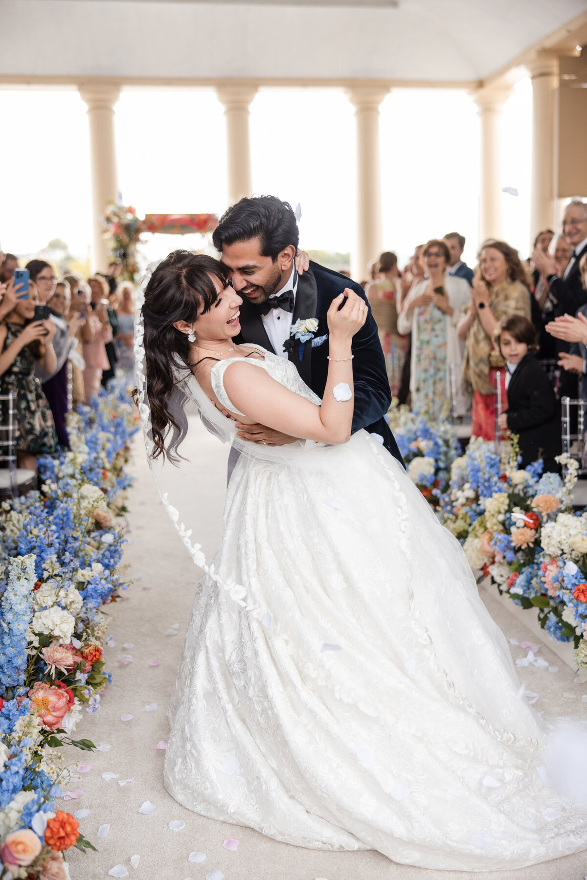 A couple shares a joyful embrace after their ceremony at Water Works in Philadelphia, surrounded by guests tossing flower petals and floral aisle arrangements in blue, peach, and white tones, framed by classical columns and natural light.