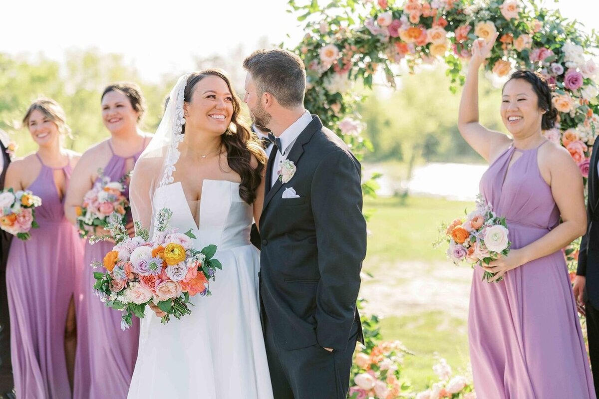 Bride and groom smiling under whimsical floral arch with bridesmaids holding pastel garden bouquets