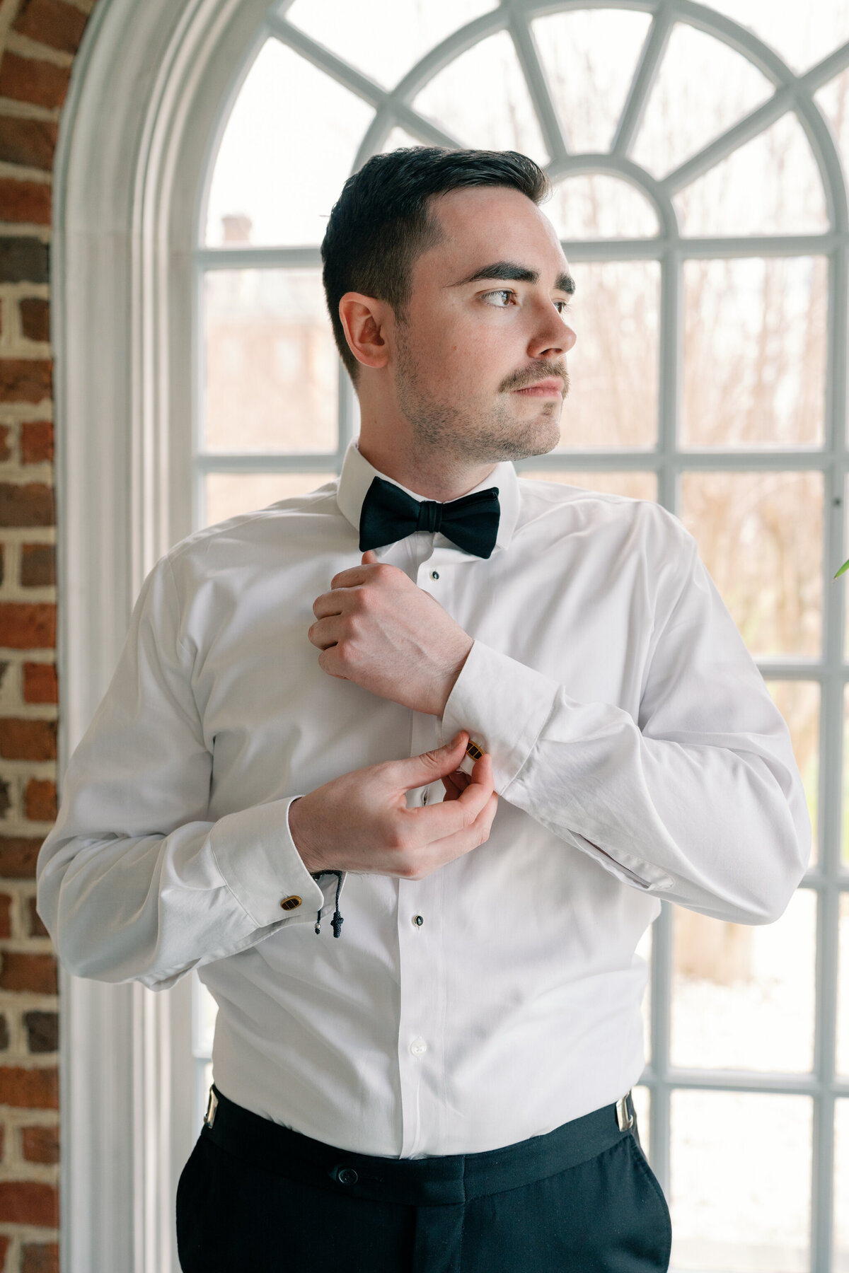 Estate at River Run Wedding in Maidens, VA | Wedding Photographer in Richmond | groom in white shirt with black pants and black bowtie adjusting cufflinks in front of an arched window at the Estate at River Run