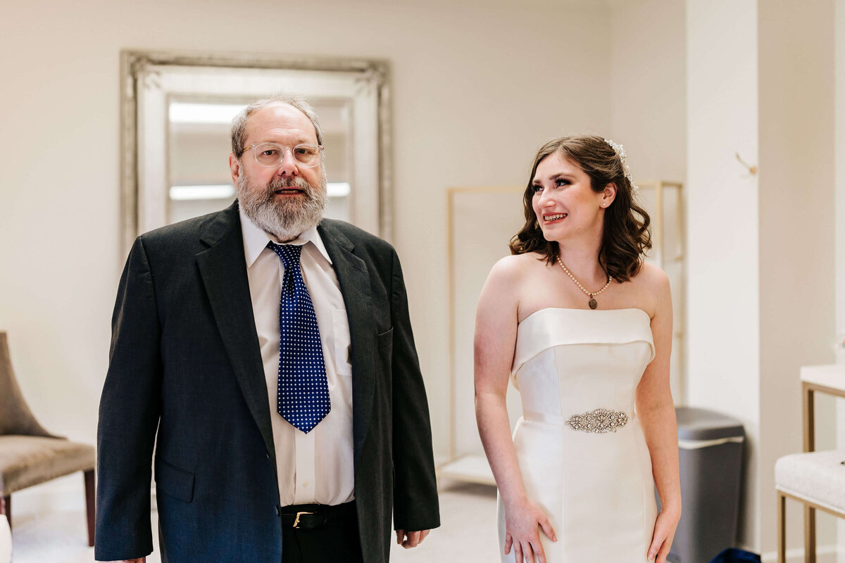 A man in a suit and tie stands next to a smiling woman in a strapless white wedding dress. They are indoors, possibly preparing for a wedding, with a mirror and chairs visible in the background.
