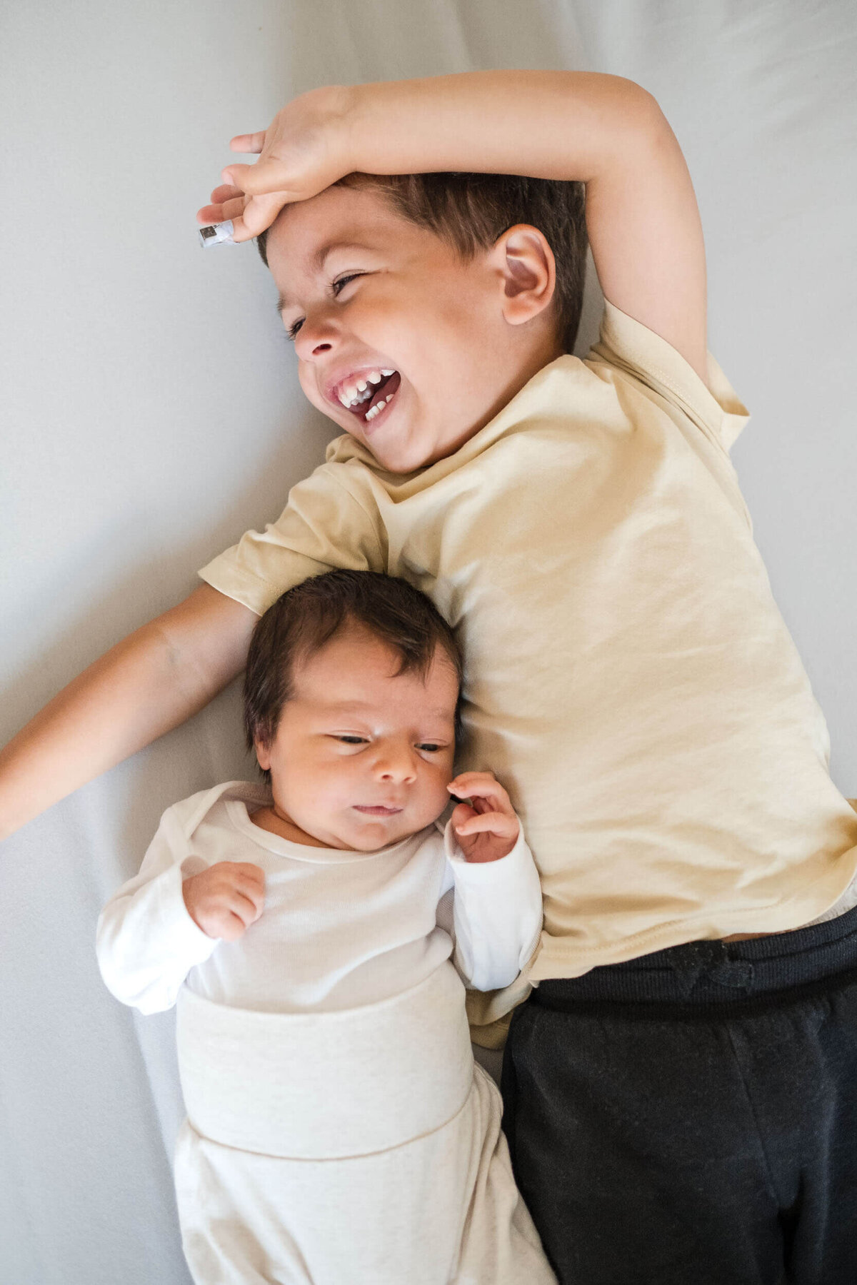 Toddler laughing while lying next to their newborn sibling on a bed