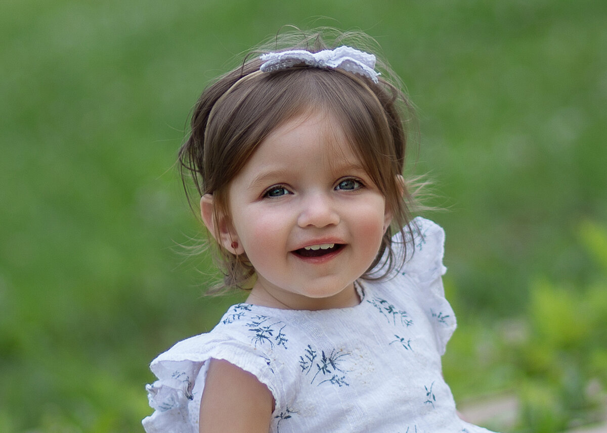 Little girl is sitting in the grass smiling at her family photoshoot in a brooklyn ny park. Captured by brooklyn family photographer Chaya Bornstein.