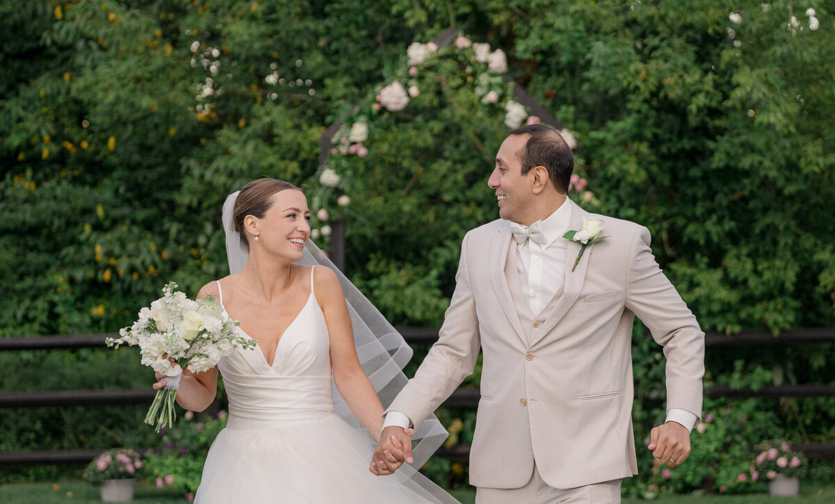 A bride with white flowers and a groom in a tan suit run by a wooded area
