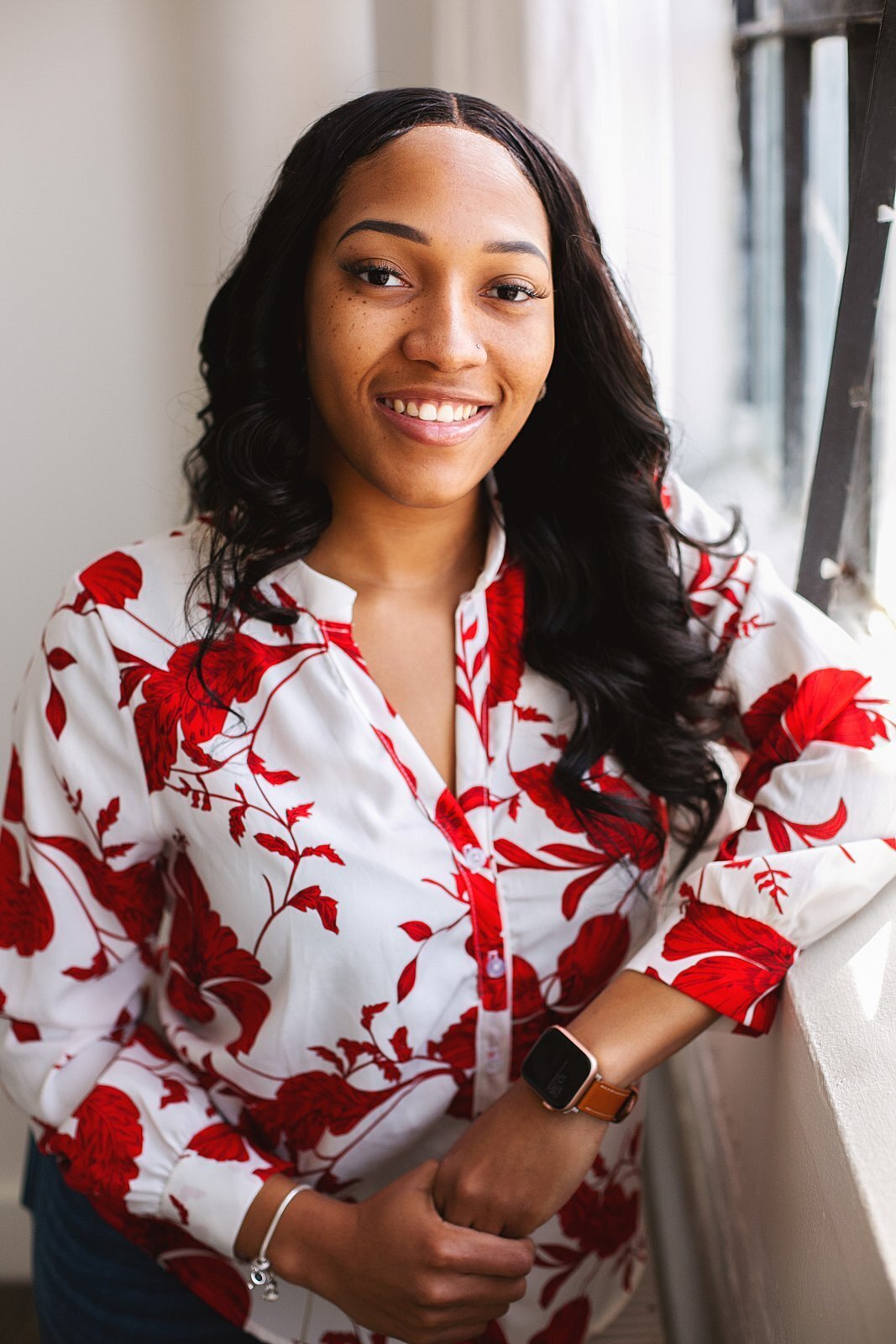 a-young-woman-in-a-red-and-white-floral-blouse-smiling-brightly-by-a-window-for-a-corporate-headshot-in-downtown-indianapolis