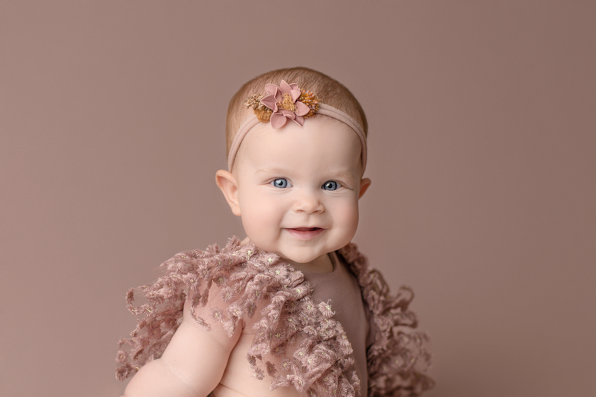 Close up image of baby girl dressed in mauve ruffle dress smiling at the camera on a mauve backdrop during. her 6 month sitter milestone session in Pensacola, FL