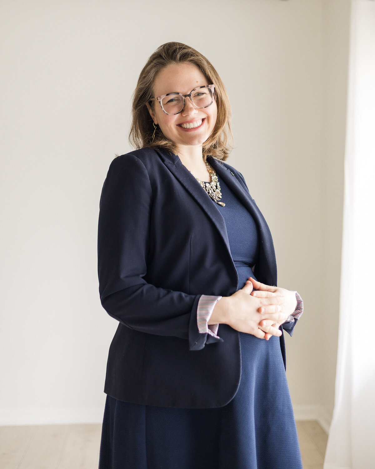 Brunette blonde pregnant woman standing with her hands over her stomach, wearing glasses, a navy dress with a navy dress jacket, smiling at the camera