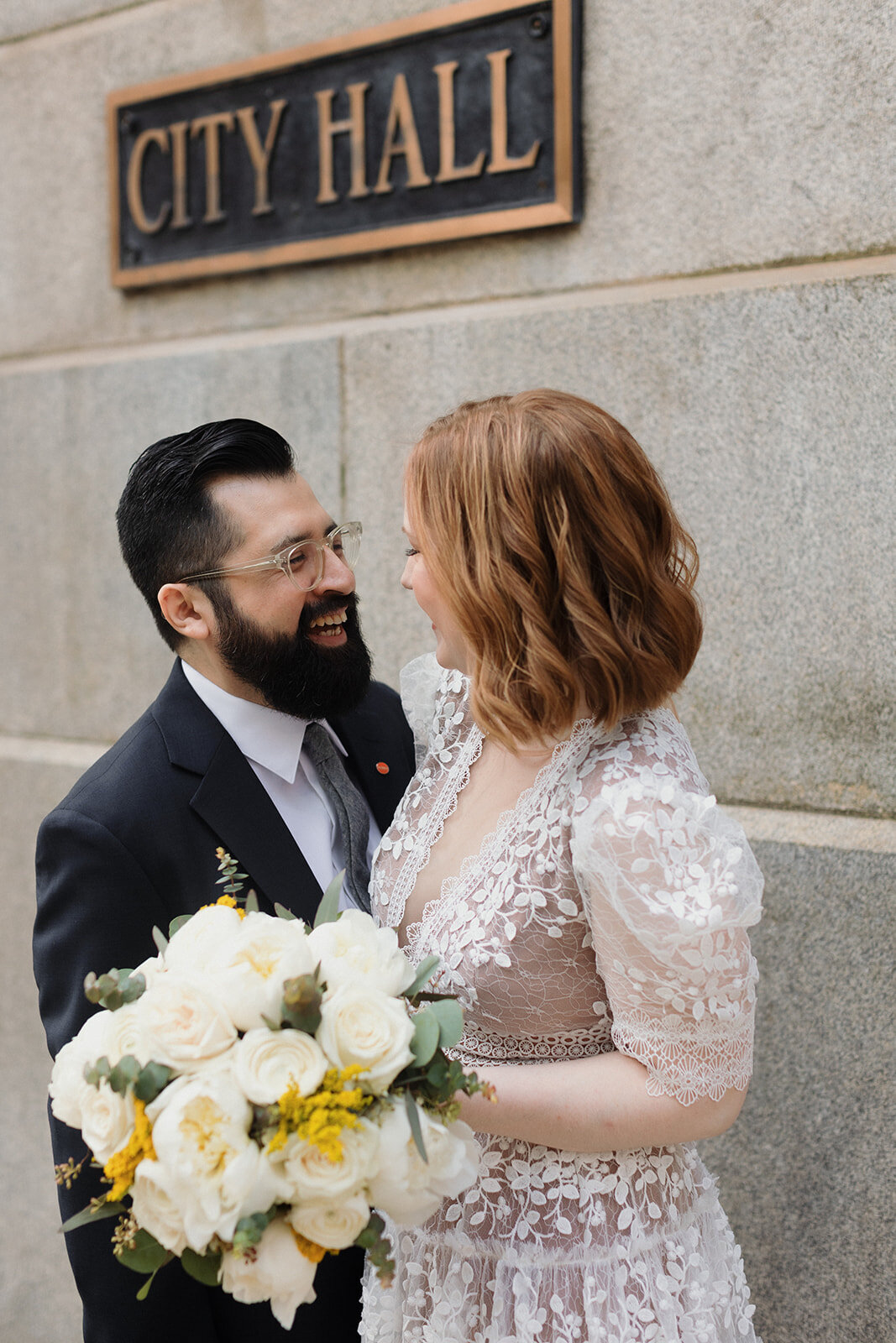 couple is standing under Chicago city hall sign after their wedding ceremony. They're smiling at each other and laughing with joy.