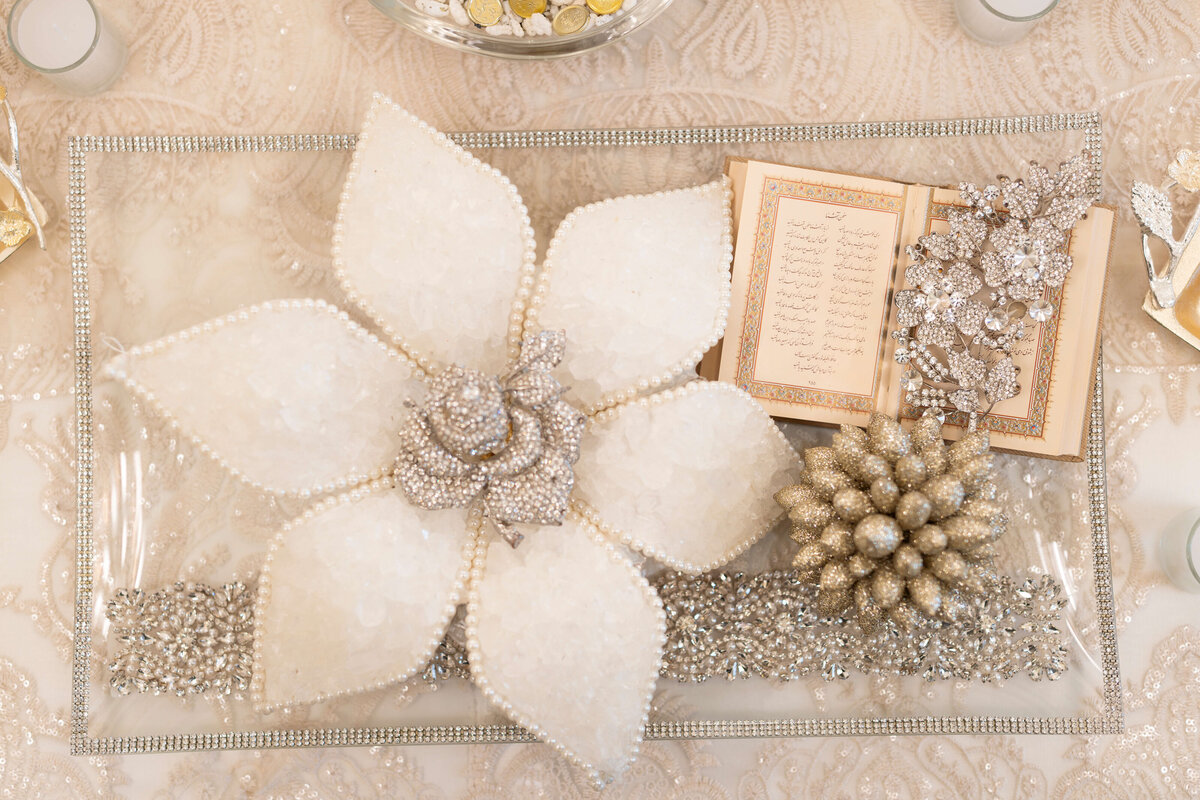 A decorative flat lay featuring a large white flower adorned with pearls and crystals, an open book with ornate script, and shimmering silver and gold embellishments on a patterned background.