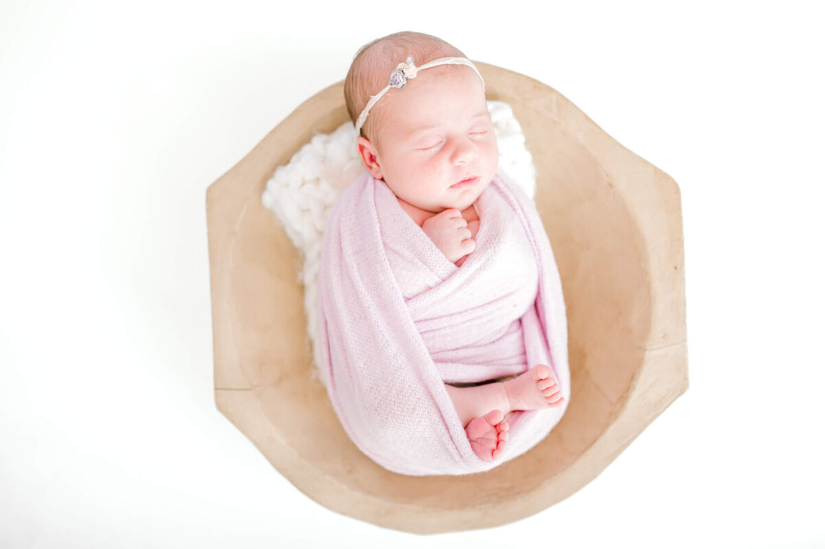 Newborn baby girl in bowl wrapped in light lilac and floral on her headband