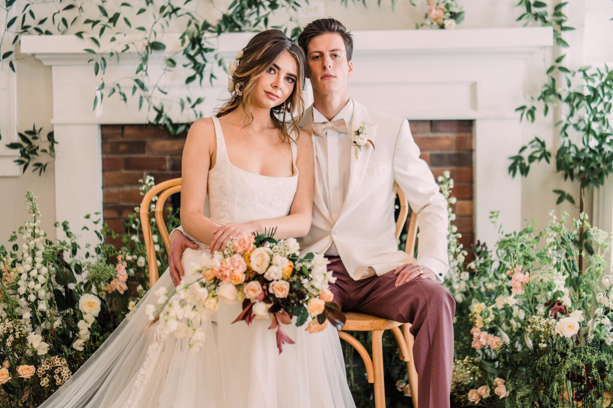 Bride wearing Alexandra Grecco gown holding bouquet & groom in white tux seated before flower & vine-covered fireplace mantle