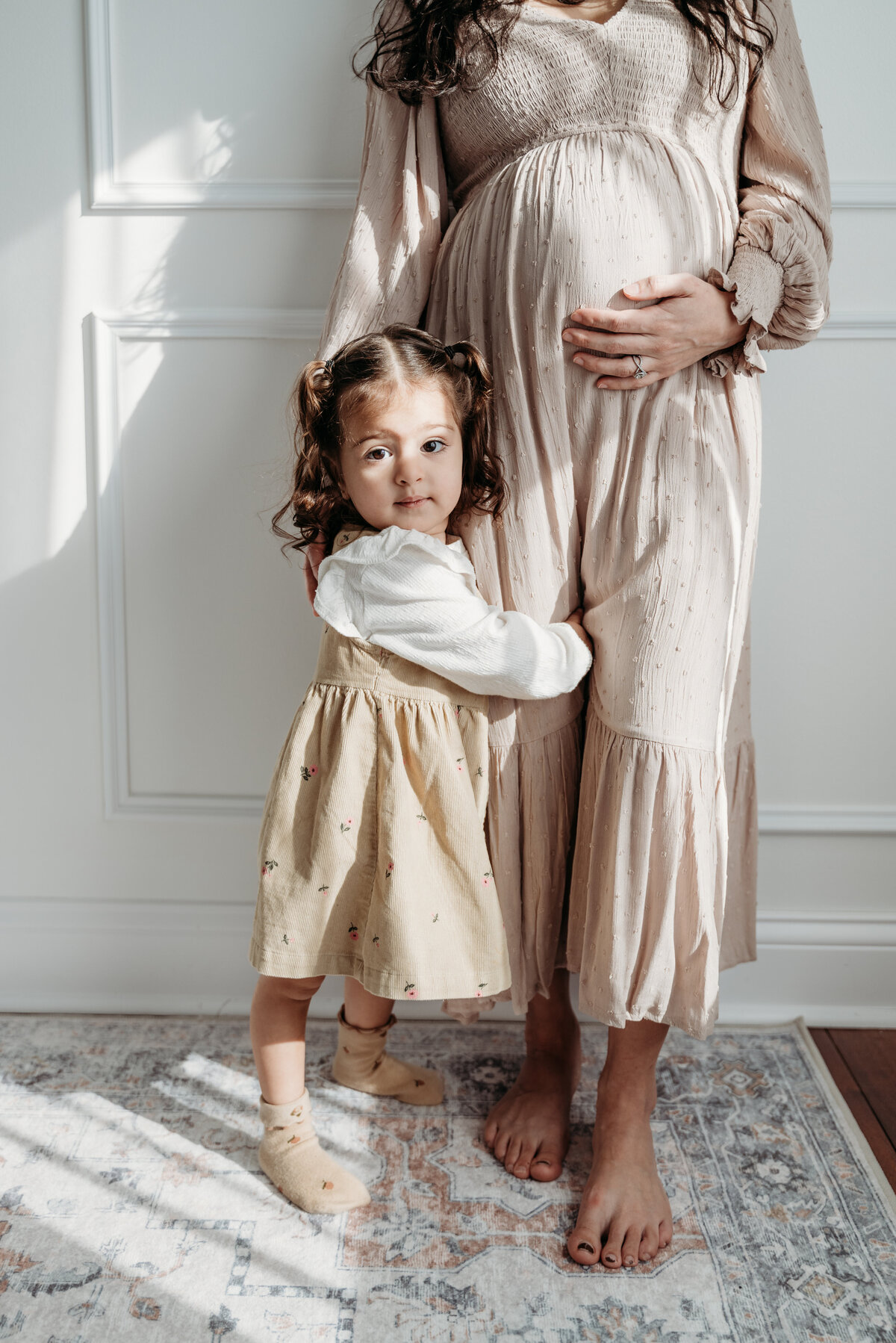 Toddler girl hugs at her moms feet as the mom embraces both her and the baby bump in an elegant, bright studio in bartlett, il