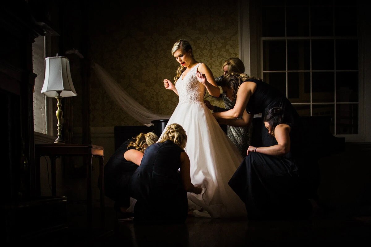 An atmospheric shot of a bride being dressed and attended to by bridesmaids, with dramatic lighting accentuating the textures of her gown.