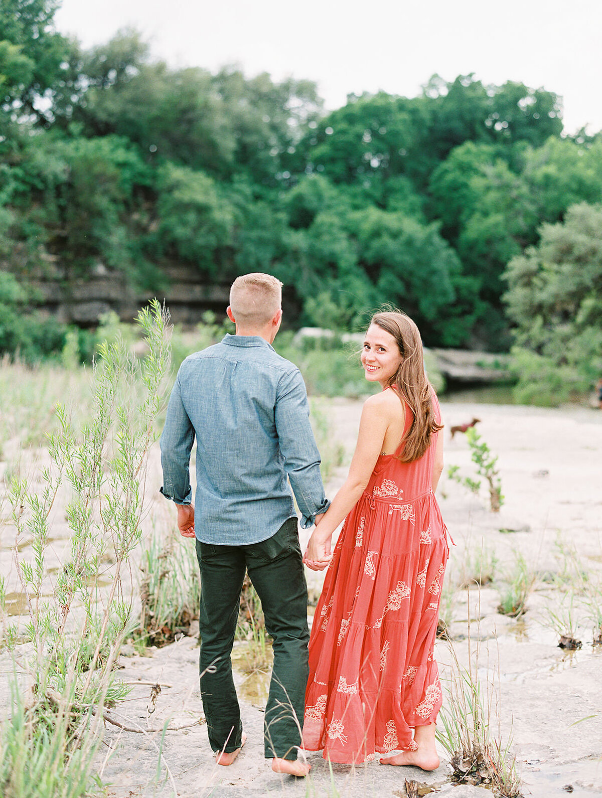girl in red dress looks over her shoulder while holding her fiance's hand