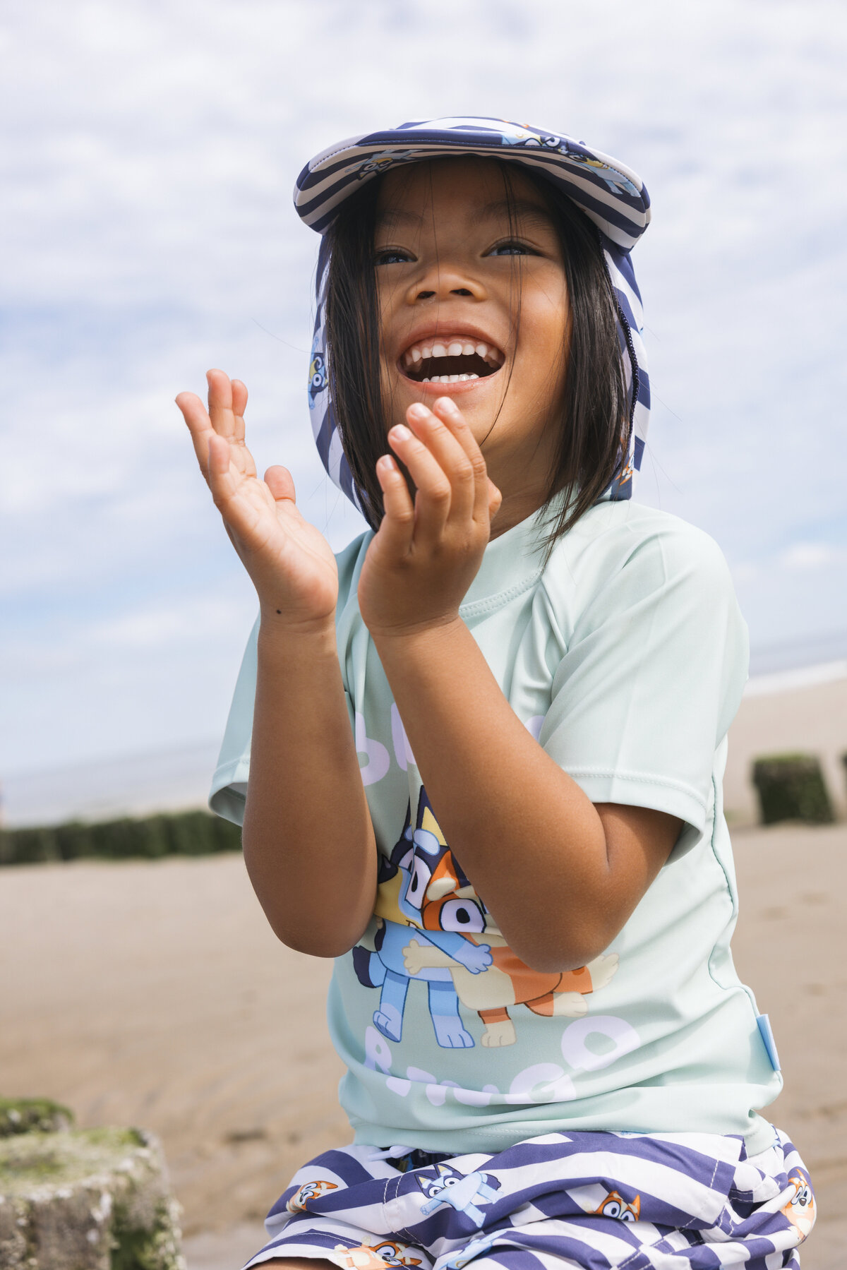Happy child clapping hands while wearing a Swim Essentials outfit with Bluey print, sitting on the beach.
