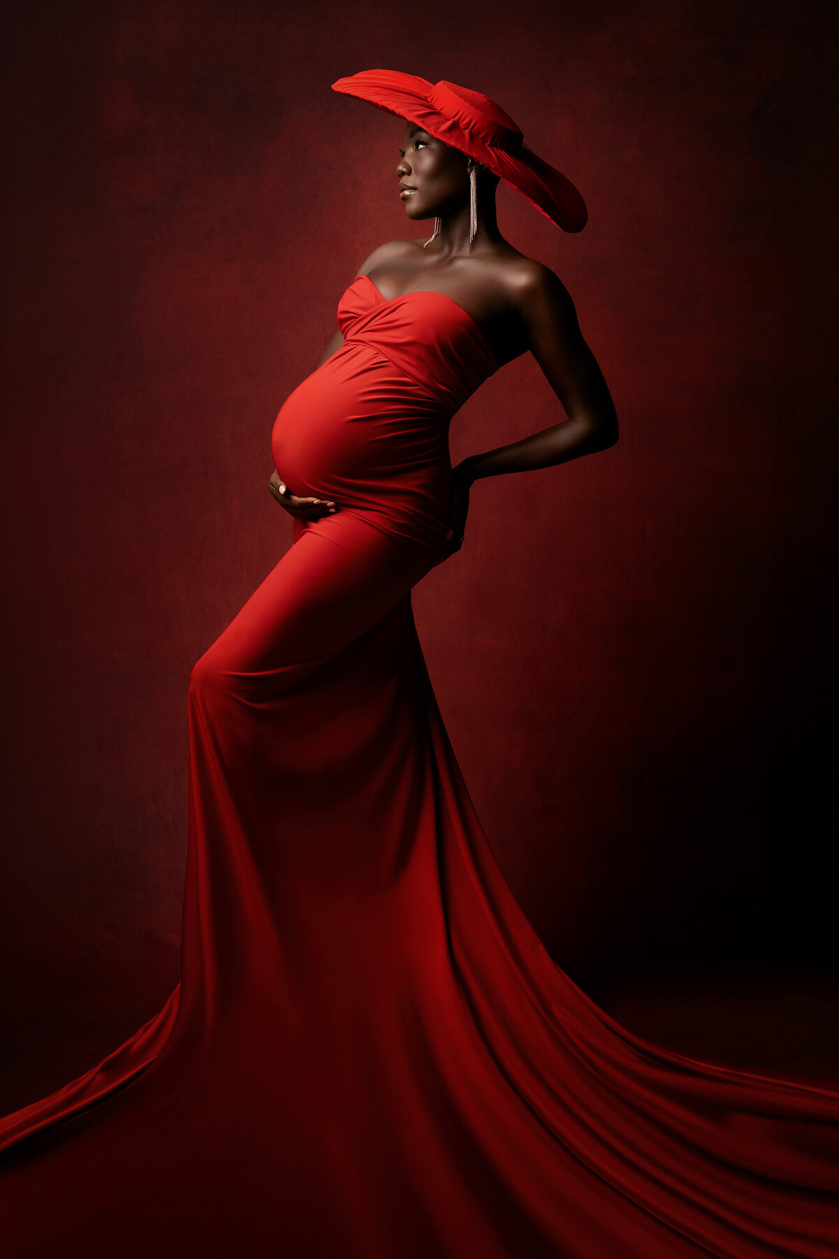 Exquisite woman in a vibrant red gown with a matching red hat on a red fine art background. Captured by DFW maternity photographer Lindsay Walden.