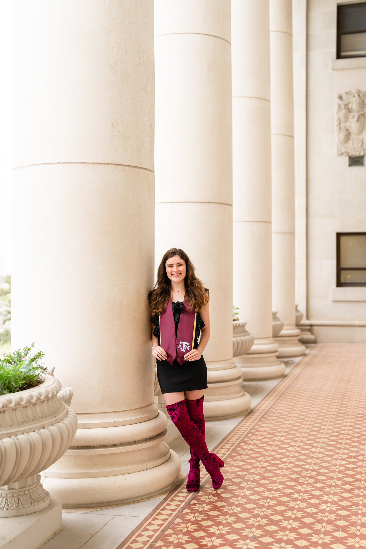 Texas A&M senior girl holding Aggie stole and smiling while leaning against column while wearing black dress and tall maroon boots at Administration Building