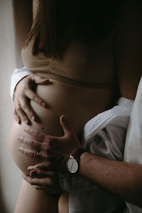 Mains posées sur le ventre rond d'une femme enceinte. Séance photo grossesse à la maison.