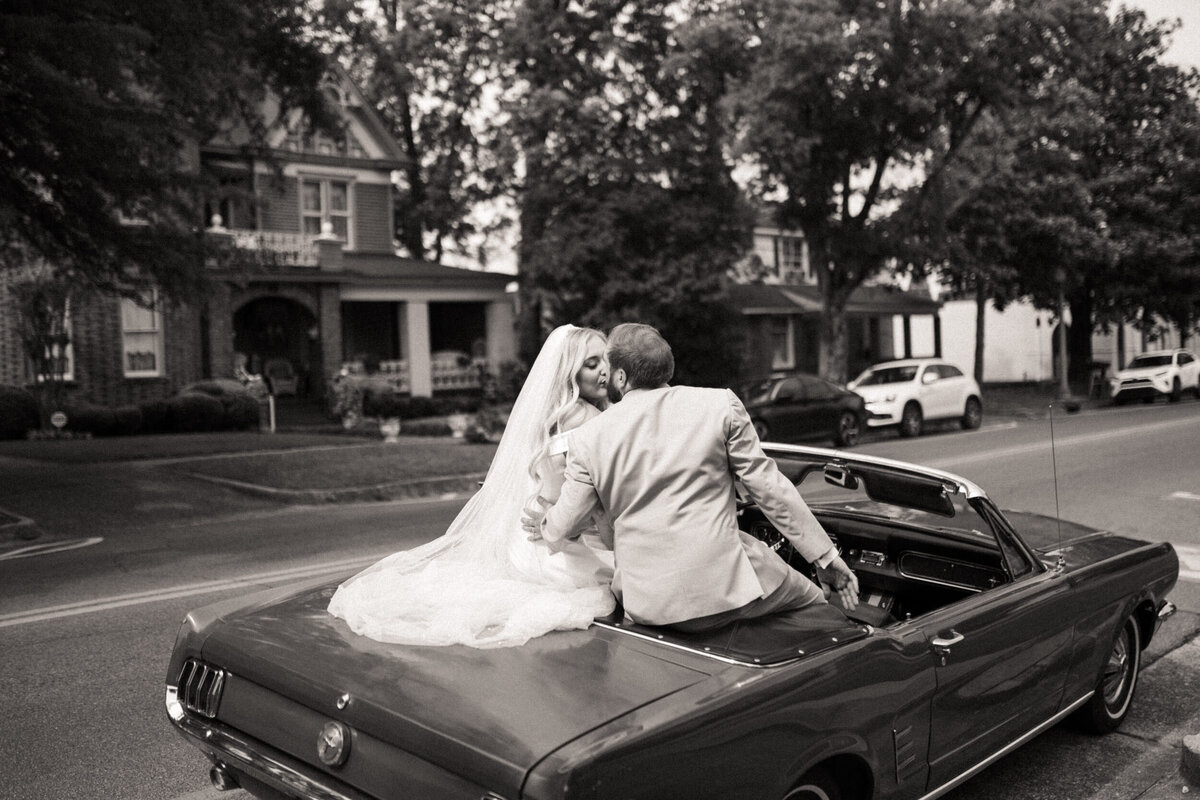 Bride and groom sitting on the back of a convertible car kissing and driving down the street