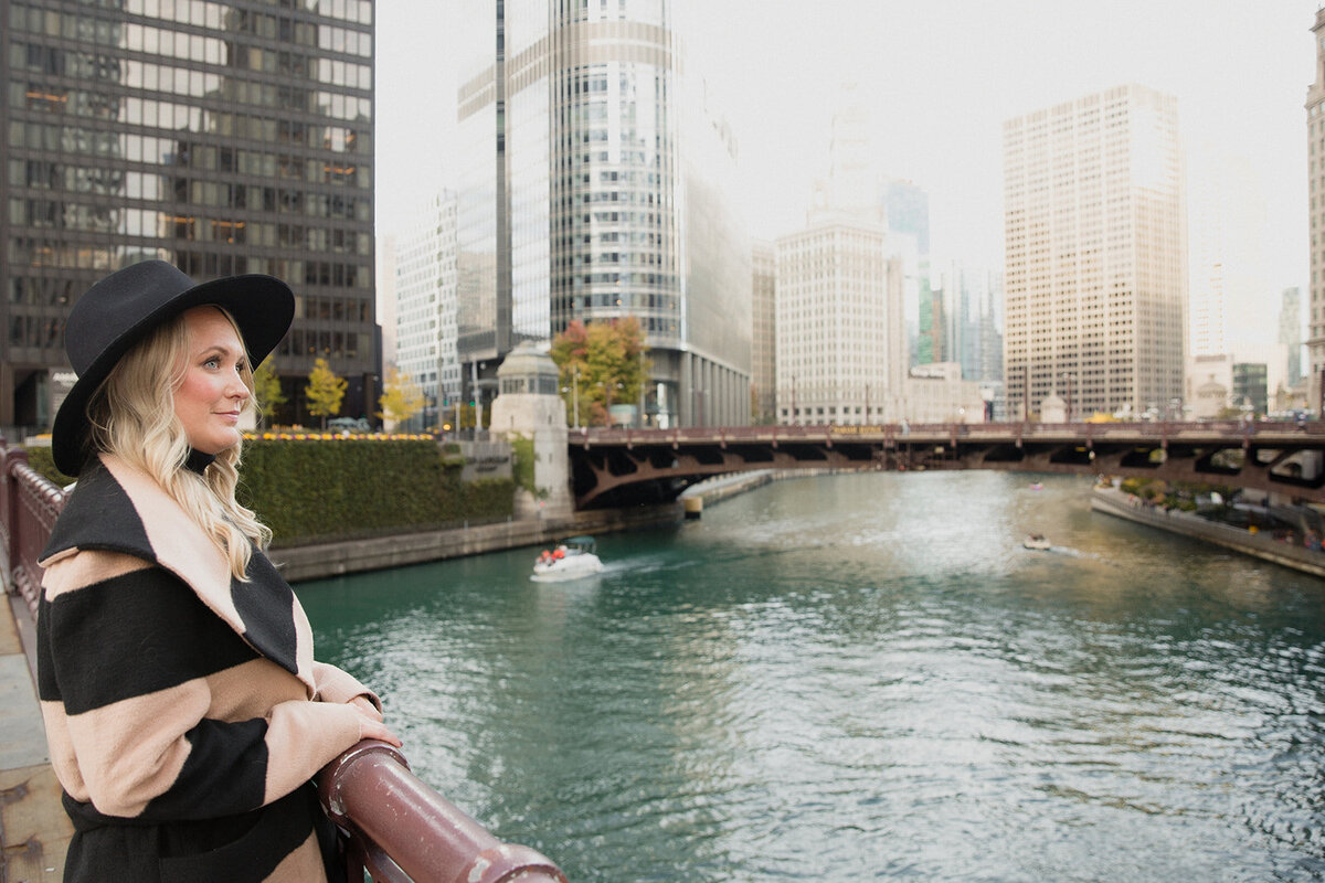 blonde woman in black and beige striped coat and black hat is standing on a bridge over the Chicago River and looking off into the distance.