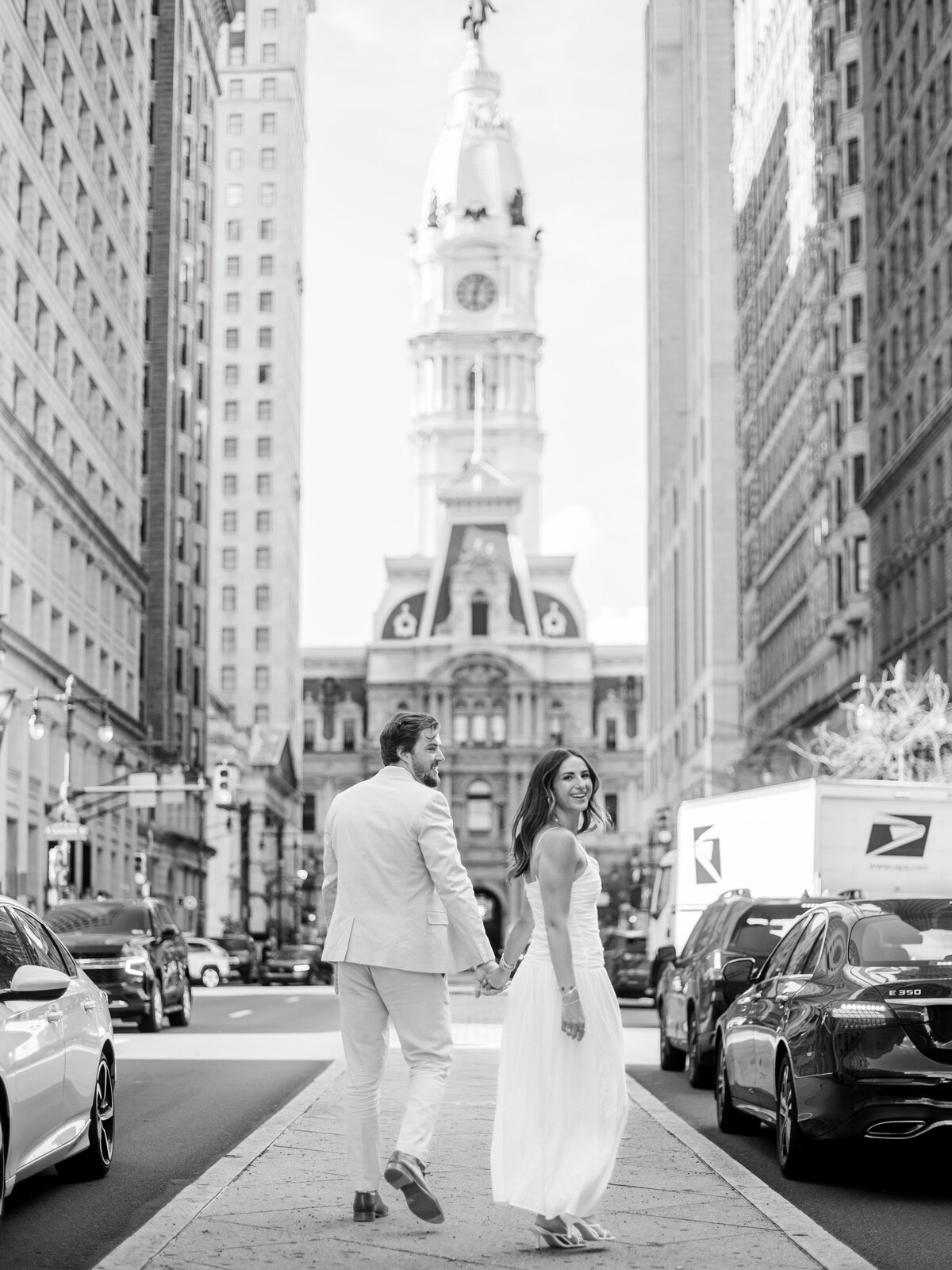A black-and-white portrait of a couple walking hand in hand down Broad Street in Philadelphia, with City Hall standing tall in the background. The woman glances back with a joyful smile while her partner looks toward her, capturing a candid moment of connection. Photographed by Elizabeth Kane Photography, a Luxury Philadelphia Wedding Photographer.