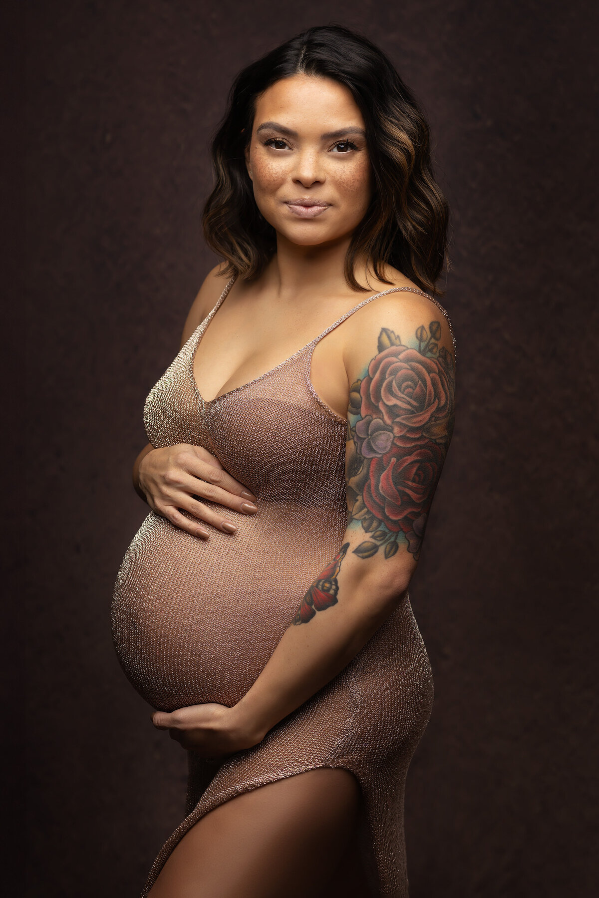 Maternity photograph of a woman in a shimmering dress, cradling her belly with a serene expression. She has a colorful floral tattoo on her arm, set against a dark background.