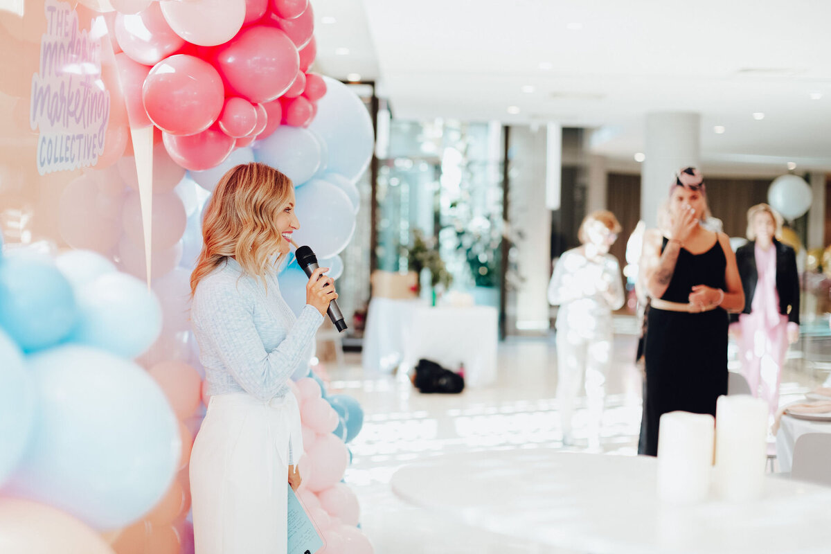 Emily, the event host, speaking at a marketing event in front of balloons.