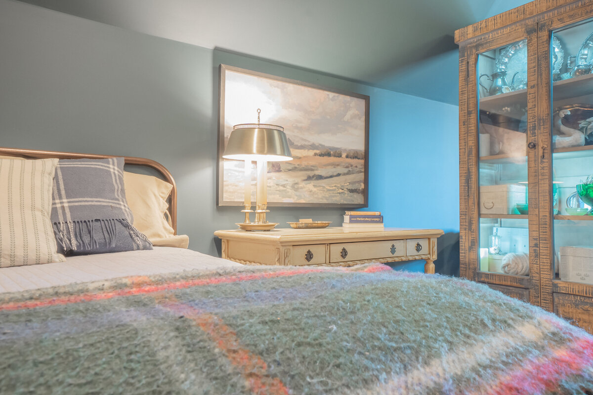 Bedroom with metal frame bed tartan plaid blanket and pillows, alongside a yellow wooden desk and wooden cabinet with glass doors