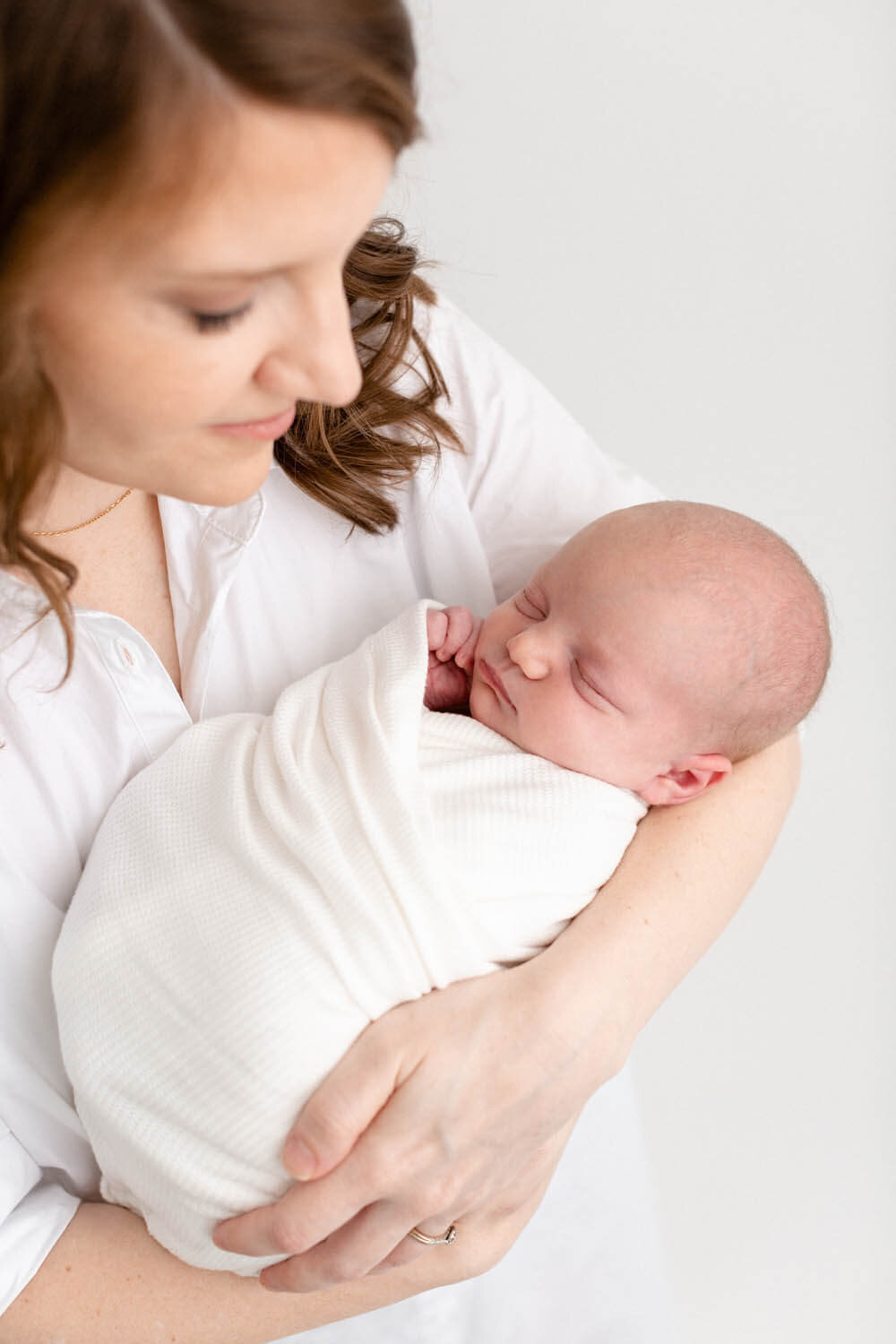 Mama in a white buttoned down collared shirt is holding her baby in her arms and looking down at her with a soft smile on her face. Baby is swaddled in white and sleeping with one hand up by her little face.