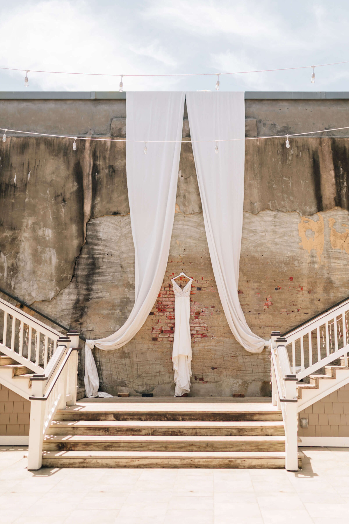 A white wedding dress hangs on a wooden hanger against a rustic brick and concrete wall, framed by flowing white drapes on either side.
