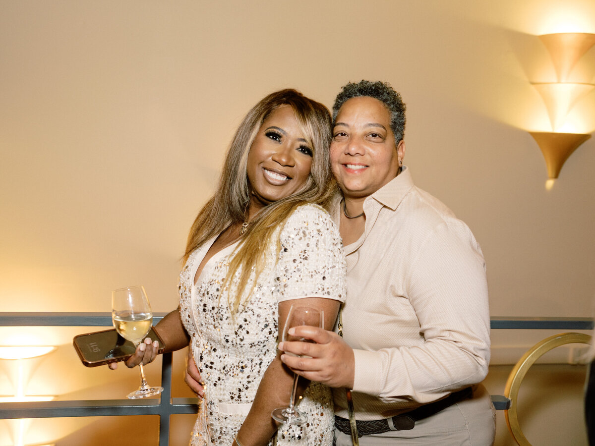 Two women embrace joy at a wedding, holding drinks and smiling radiantly, showcasing a moment of love and celebration in a festive environment.