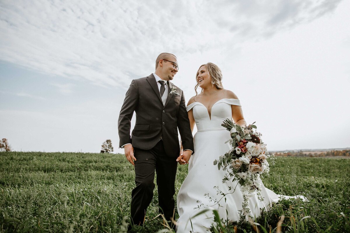 Bride and groom holding hands in Exeter, Ontario field. Bride and groom are holding hands and looking at each other. The bride is holding a large floral bouquet in the other hand.